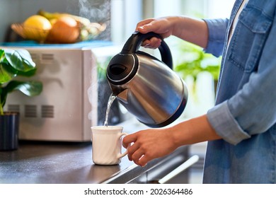 Female hands pouring boiling water from a modern metal stainless kettle in a glass cup for brewing tea in the kitchen at home