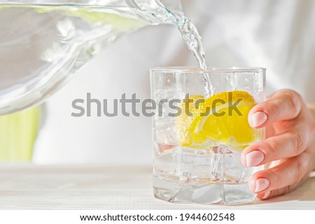 Female hands pouring water from the decanter into a glass beaker with lemon and ice. Health and diet concept. Quenching thirst on a hot day.