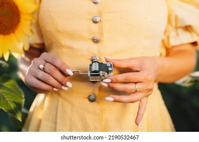 Female Hands Rotating Gears Of Old Music Box Mechanism. Lady Turning The Lever Of Retro Small Metallic Carillon. Woman In Sunflowers Field Listening To Music Which Playing. High Quality Photo