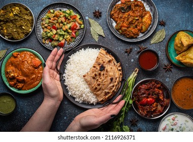 Female Hands Serving Indian Ethnic Food Buffet On Rustic Concrete Table From Above: Curry, Fried Samosa, Rice Biryani, Dal, Paneer, Chapatti, Naan, Chicken Tikka Masala, Dishes Of India For Dinner