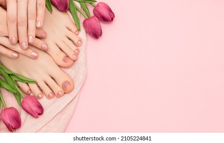 Female hands with spring nail design. Glitter pink nail polish pedicure. Female hands and feet with tulip flowers on pink fabric background with copy space.