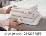Female hands with stack of clean bed sheets and cotton flowers on bench at home, closeup