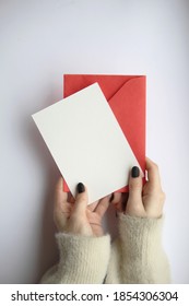 Female Hands In A Sweater With Manicure, Holding A White Sheet And A Red Envelope On A White Background. Postcard Layout