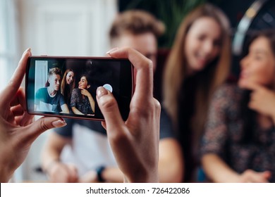 Female hands taking photo with smartphone of young cheerful friends, blurred background