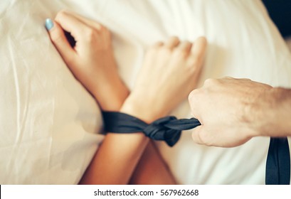 Женские руки, связанные галстуком черного мужчины, натянутым мужской рукой на кровати с белыми простынями - Женщина лежит на кровати со связанными руками лентой - Ролевая игра пары, занимающейся сексом в спальне