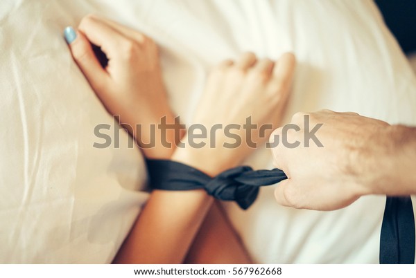 Female hands tied with a black mans tie pulled by
a man's hand on a bed with white sheets - Woman laying on a bed
with bound hands with a ribbon - Role playing game of couple having
sex in the bedroom
