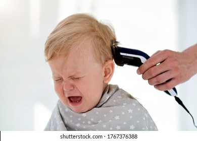 Female hands trim a crying baby with an electric hair clipper in a hairdresser. First haircut. Mom cuts boy is home during quarantine