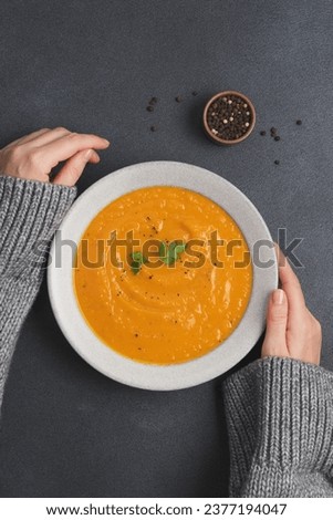 Female hands in warm sweater holding pumpkin soup in bowl on black background. Eating traditional seasonal food