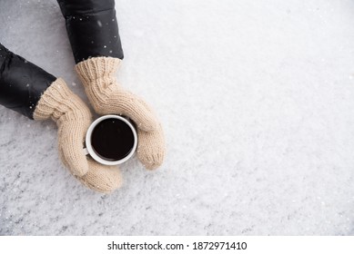 Female Hands In Woolen Knitted Gloves Holding Cup Of Warm Black Coffee Outdoors On Snowy Background. Top View Of Hands And Coffee Mug. Happy, Frosty Winter Morning