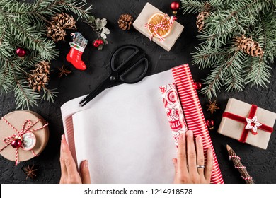 Female Hands Wrapping A Christmas Gift On Holiday Wrapping On Black Background With Fir Branches, Red Decorations. Xmas And Happy New Year Card. Flat Lay, Top View