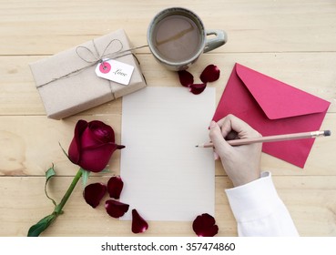 Female hands writing down on letter paper for Valentine's day with gift box and cup of coffee on desk, top view