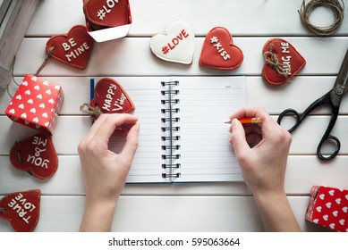 Female hands writing or drawing sketches in open notebook on white table. Bird eyes view. Gingerbread in form of heart.St. Valentines Day concept.