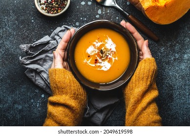 Female hands in yellow knitted sweater holding a bowl with pumpkin cream soup on dark stone background with spoon decorated with cut fresh pumpkin, top view. Autumn cozy dinner concept 