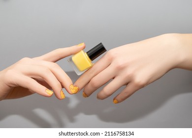 Female Hands With Yellow Nail Design. Glitter Yellow Nail Polish Manicure With Nail Art. Woman Hands Hold Yellow Nail Polish Bottle On Gray Background.