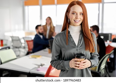 female having break time holding cup of coffee, redhead lady is looking at camera in the office, in formal wear