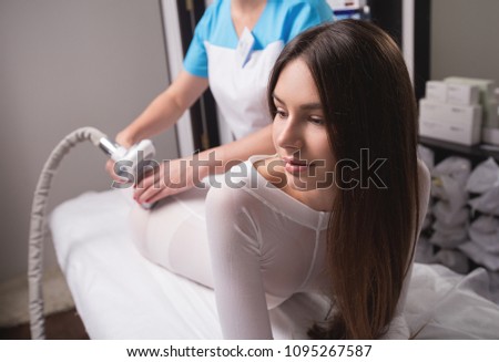 Female having procedure of massage on legs in apparatus cosmetology clinic. Woman in special white suit getting anti cellulite massage in a spa salon. LPG treatment