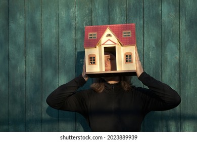 Female Head Inside Tiny Home. Woman Holding Toy House In Front Of Her Face, Her Eye Looks Through Open Door. Real Estate Concept, Isolation And Loneliness Psychology.