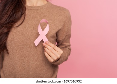 Female health check awareness. Closeup of young woman in pullover holding pink ribbon, symbol of breast cancer, timely diagnosis, support of oncology patients. studio shot isolated on pink background - Shutterstock ID 1697510932