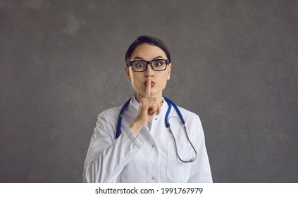 Female healthcare professional or doctor puts her index finger to her lips demanding silence. Studio portrait of a doctor on a gray background. Concept of silence and medical secrecy. Banner. - Shutterstock ID 1991767979