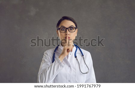 Female healthcare professional or doctor puts her index finger to her lips demanding silence. Studio portrait of a doctor on a gray background. Concept of silence and medical secrecy. Banner.