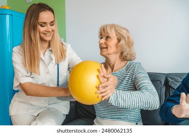 A female healthcare worker assists an elderly woman with a yellow exercise ball during a physical therapy session in a home setting. - Powered by Shutterstock