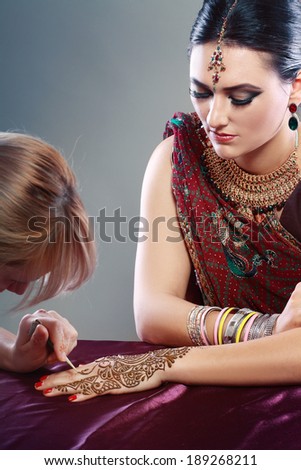 A female henna artist applies a henna design to the back of a woman's hands.
