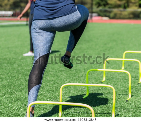 A female high school athlete performs running\
drills over yellow mini banana hurdles on a turf field with no\
shoes on, only socks in blue\
spandex.