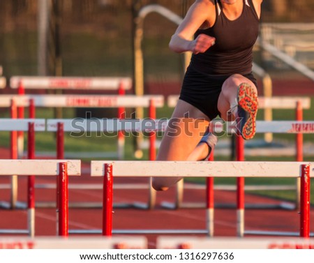 A female high school runner wearing a black uniform is racing the 110 meter high hurdles outdoors.