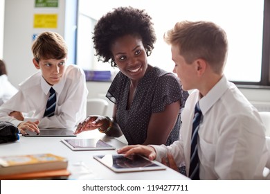 Female High School Teacher Sitting At Table With Students Wearing Uniform Using Digital Tablets In Lesson