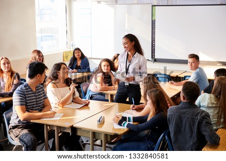 Female High School Teacher Standing By Student Table Teaching Lesson