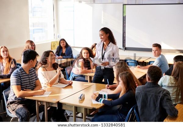 Female High School Teacher Standing By Student\
Table Teaching Lesson