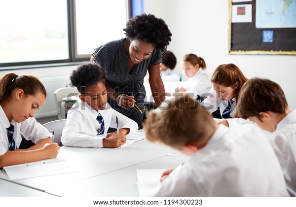 Female High School Tutor Helping\
Students Wearing Uniform Seated Around Tables In\
Lesson