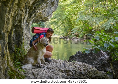 Female hiker with backpack and a golden retriever staying on a rock close to a river. in a green forest.