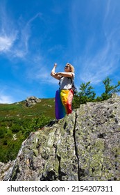 A Female Hiker With A Backpack, Perched On A Rock At The Top Of A Mountain, With A Rainbow Flag Hanging From Her Waist And Making A Sleeve Cut. With A Blue Sky And Mountainside In The Background.