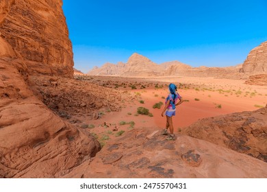 Female hiker with a colorful backpack stands on a rocky overlook at Lawrence of Arabia house amidst, gazing at the vast, serene expanse of Wadi Rum's red desert landscape under a clear sky in Jordan - Powered by Shutterstock