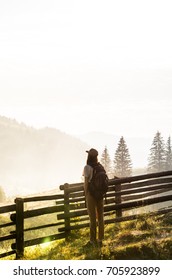 Female hiker enjoys beautiful mountain scenery at dawn in carpathian rural area. Young backpacker tourist girl stands by the fence and looks at hills with forests and mist at sunrise - Shutterstock ID 705923899