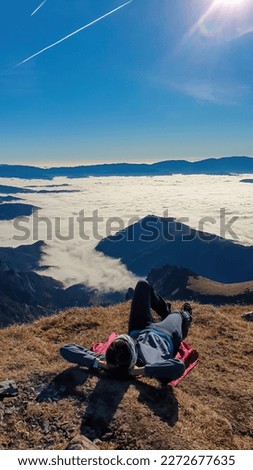 A female hiker having a break on the summit of Eisenerzer Reichenstein in Styria, Austria, Europe. The Ennstal valley is covered in clouds and fog. Hiking trail, Wanderlust, Season. Sunny day. Freedom