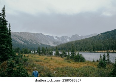 Female hiker on intimate outdoor hiking trail in the fall surrounded by tall green evergreen trees, long lake calm waters, and snow patched mountains under cloudy sky - Powered by Shutterstock