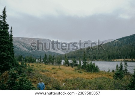 Female hiker on intimate outdoor hiking trail in the fall surrounded by tall green evergreen trees, long lake calm waters, and snow patched mountains under cloudy sky
