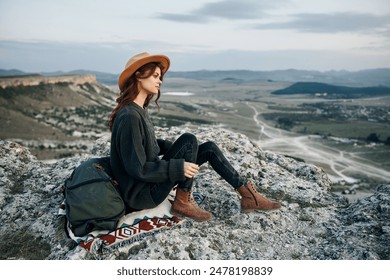 female hiker resting on rocky summit with backpack and hat, overlooking vast mountain landscape - Powered by Shutterstock