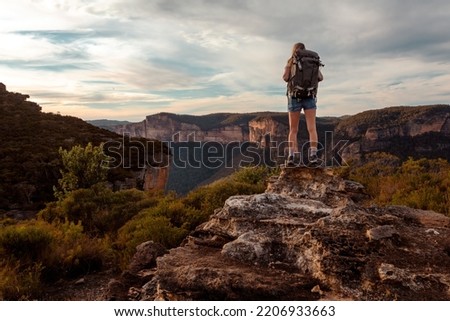 Female hiker stands on a rock carrying a large backpack ... beyond her is views to mountains with sheer cliffs  and valley
