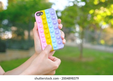 Female holding bright pop it bauble smartphone case infront of green garden. Antistress silicone toy. Simple dimple. Fidget gadget. Autism awareness. Stress reliever
