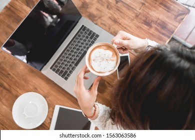 Female Holding Coffee Cup In Hands Computer And Cellphone  On Table At Home, Top View. Young Woman Drinking Coffee With Laptop, Mobile Phone And Tablet.
Woman Freelancer Work From Home WFH Concept.