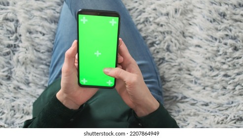 Female Holds Smartphone With Green Screen In Hands. Top View, Zoom In Close Up, Living Room, Sofa. Using The App, Swipe Up. Unrecognizable Woman.