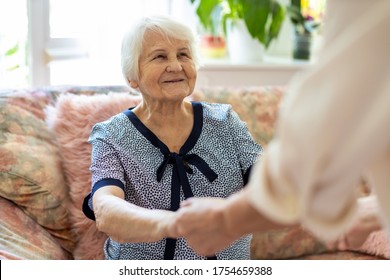 Female Home Carer Supporting Old Woman To Stand Up From The Sofa At Care Home