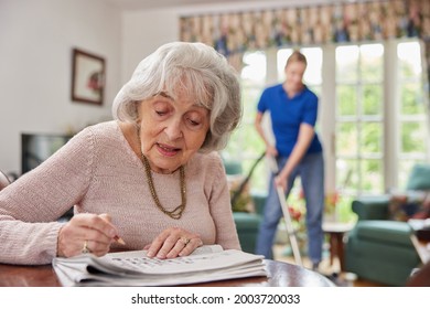 Female Home Help Cleaning House With Vacuum Cleaner Whilst Senior Woman Does Crossword In Newspaper