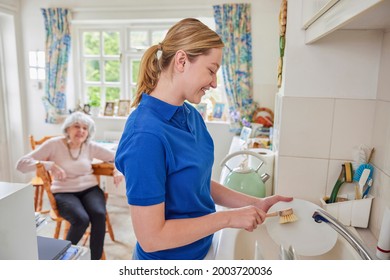 Female Home Help Cleaning House Doing Washing Up In Kitchen Whilst Chatting With Senior Woman 