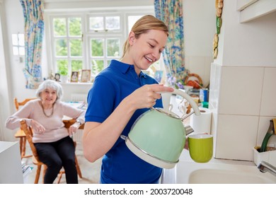 Female Home Help Making Cup Of Tea In Kitchen Whilst Chatting With Senior Woman 