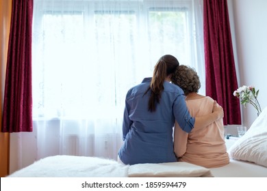 Female Home Nurse Hugging Elderly Woman On Bed. Back View Of Female Nurse With Her Arm Around Elderly Patient Shoulder.