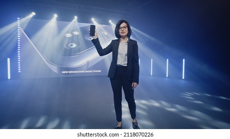 Female host in a suit on stage showing a new fashionable device, during the presentation of the release near the LED screen with a 3D mock-up in a large illuminated room